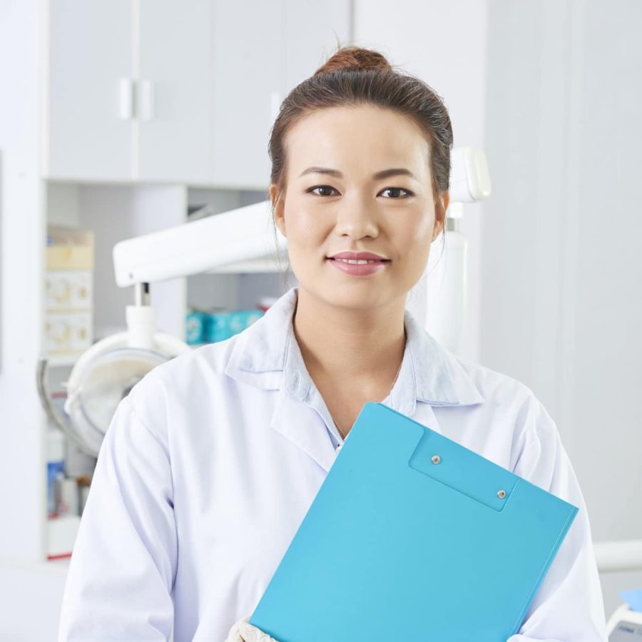 Vertical medium portrait of beautiful Asian female dentist holding blue clipboard looking at camera smiling