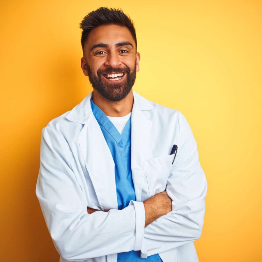Young indian doctor man standing over isolated yellow background happy face smiling with crossed arms looking at the camera. Positive person.