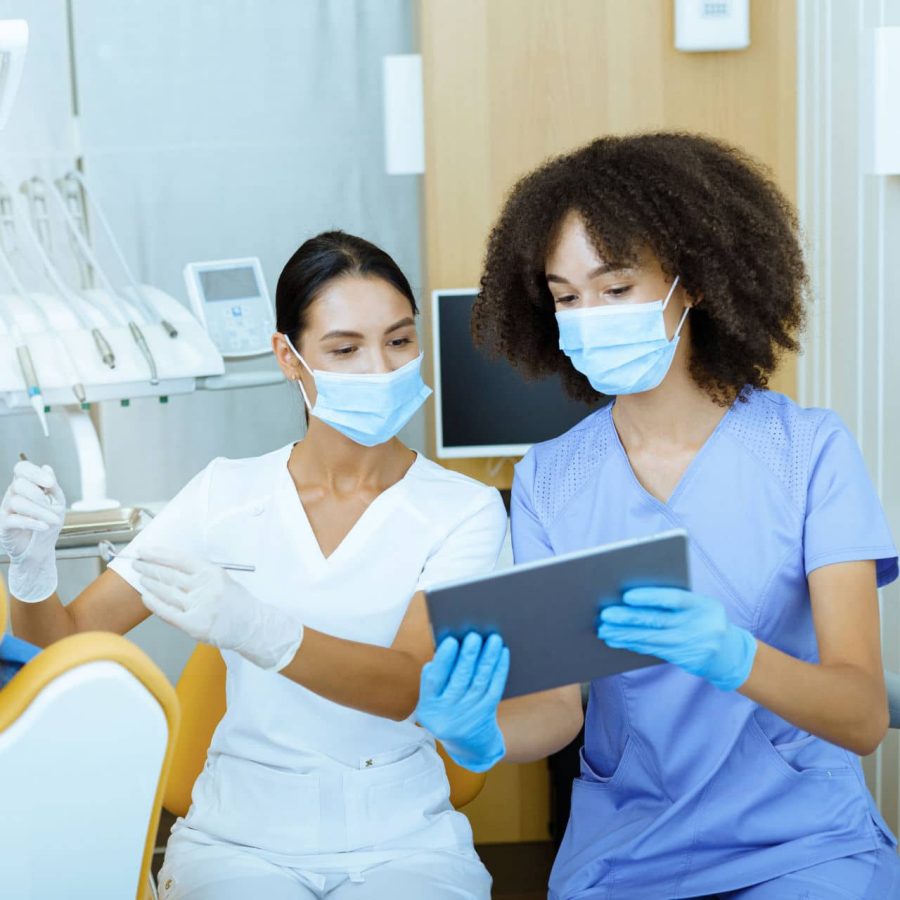 Modern technology and X-rays in dental office. Young european and african american female doctors in protective masks and rubber gloves, treat child in chair and look at tablet in clinic with tools